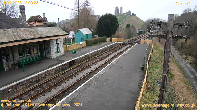 A view of Corfe Castle railway station featuring a stone platform with a green bench and a shelter. The tracks run parallel to the platform, leading into the distance. On the left, there is a wooden fence and a small green sign labeled "WAY OUT." Behind the station, a hill rises with the ruins of Corfe Castle visible at the top. A telephone pole with wires stands on the right side of the image, and there are scattered trees and shrubs in the background. The sky is overcast.