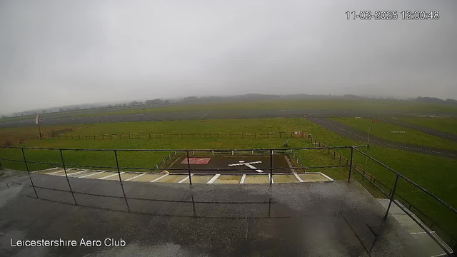 A cloudy and overcast scene showing a grassy area and an airstrip. In the foreground, there is a railing with a view of a marked landing area featuring a white 'X' shape on the ground. In the background, the airstrip is visible, leading towards a horizon obscured by clouds. The image is taken from a raised viewpoint, possibly from a clubhouse or control tower of an aero club. The date and time are displayed in the top right corner.