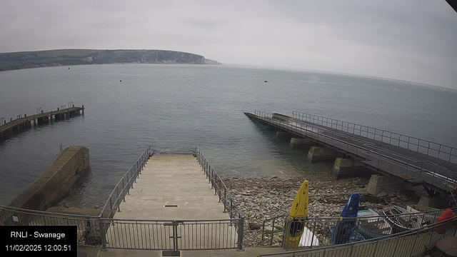 A view of a calm sea with a cloudy sky, taken from a webcam. In the foreground, there are two piers extending into the water. The left pier is made of wood and has several posts, while the right pier has a slight incline and a railing along its side. At the base of the piers, there are rocky shores. On the right side, two kayaks are resting on the shore; one is yellow and the other is blue. The distant shoreline features cliffs covered with greenery. The time and date displayed at the bottom left indicate it is noon on November 2, 2025.