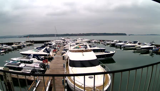 A view of a marina on a cloudy day, featuring numerous boats docked in the water. In the foreground, a large white yacht with a black cover sits prominently. Surrounding it are various smaller boats and yachts, some with blue and white colors. The water appears still with reflections of the boats visible. In the background, distant hills are faintly visible against the overcast sky, which is gray with a soft light.