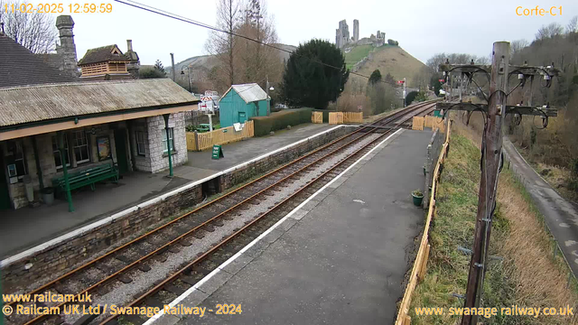 A view of a railway station platform. To the left, there is a building with a sloped roof and a few windows, alongside a green bench. The platform is made of grey concrete and has a track running alongside it with visible wooden rail ties. In the background, an old castle ruin sits atop a green hill, with trees surrounding the area. A wooden fence lines the right side of the view, and a telephone pole with wires is positioned on the right. The sky appears cloudy.