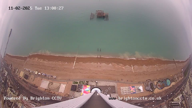 A panoramic view from a high vantage point overlooking a beach. The sandy shoreline stretches along the bottom of the image, with gentle waves lapping at the shore. In the foreground, there are several beach umbrellas and a coastal path, with a few small structures visible on the sand. Out in the water, an old pier is partially submerged. The ocean appears calm, reflecting shades of blue and green, while the sky is overcast. The bottom of the image includes a circular structure, possibly an entrance or observation deck, leading down from the camera's position.