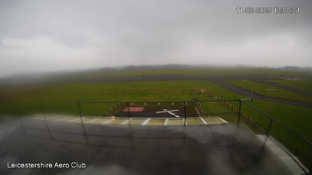 A view from a low vantage point at an airfield on a foggy day. The foreground shows a helipad marked with a white "H" on a red background, surrounded by a fenced area. The grass is green and wet, reflecting the dull grey sky filled with thick clouds. In the distance, the runway is visible, bordered by grass and low fencing, leading off into the foggy horizon. The image displays a gloomy atmosphere with limited visibility.