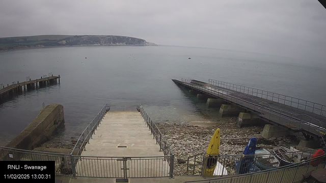 A cloudy day at a coastal area featuring a calm sea. To the left, there is a wooden pier extending into the water. In the foreground, a stone ramp leads down to the shoreline, bordered by railings. Below the ramp, rocky textures are visible, alongside a few small boats in varied colors—yellow, blue, and red—secured near the water's edge. The distant shoreline is partially obscured by clouds.