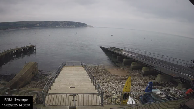 A calm coastal scene features a smooth, reflecting body of water under a cloudy sky. In the foreground, there are steps leading down to the water, bordered by a railing. To the left, a wooden pier extends out over the water, and there are several colorful kayaks—yellow, blue, and red—secured nearby. The background reveals a rocky shoreline and a sloping green hillside. Some small boats can be seen floating on the water.