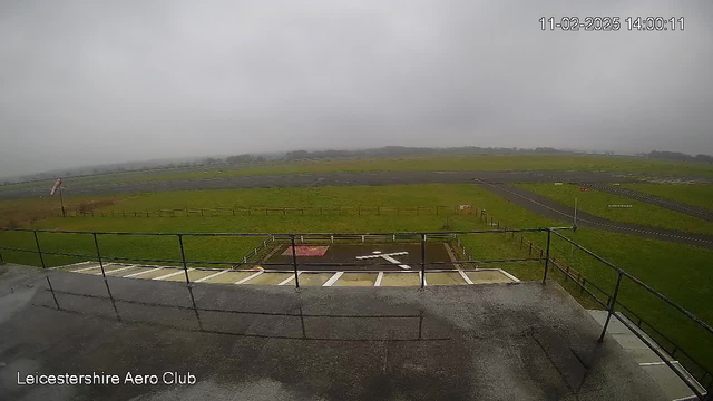 A cloudy day at Leicestershire Aero Club, showing a wide grassy field and runways. In the foreground, a low railing encloses a concrete balcony overlooking a helipad marked with a white cross. The background features an expansive green landscape fading into the mist. A windsock is visible, indicating wind direction. The time displayed is 14:00 on February 11, 2025.