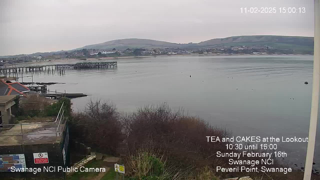 A gray, overcast sky looms over a calm body of water reflecting the dull light. In the foreground, there is a coastline with some shrubs and grass. To the left, a wooden pier extends into the water, with several pilings visible. Numerous small buoys float in the water, and in the distance, buildings line the shore along with gentle hills rising behind them. The image includes text indicating the location as Swanage NCI, the date, and details of a tea and cakes event.