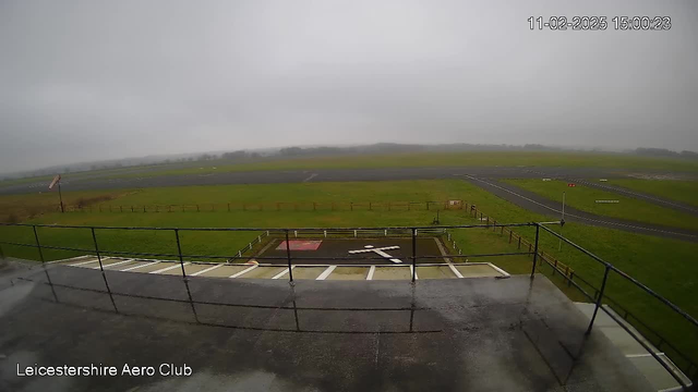 A gray and cloudy sky is above an open field with a runway. The ground is wet, indicating recent rain. In the foreground, there is a railing overlooking the field. A white helipad marked with a large cross is visible, along with a red square area beside it. In the distance, there are some trees and a small structure. The scene is quiet and desaturated due to the overcast weather.