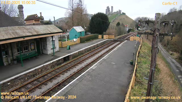 A view of a railway station with a stone building featuring a sloped roof and wooden overhang. A green bench is positioned on the platform. In the background, a small teal-colored shed is visible alongside a wooden fence with a "Way Out" sign. Train tracks run alongside the platform, leading off to the right where an area of grass and trees can be seen. A hill with ruins is in the distance, with a cloudy sky above. The scene is set in a rural environment.