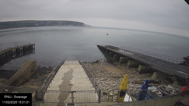 A view of a rocky shoreline with a concrete ramp leading down to the water. There is a small wooden pier on the left side extending into the calm water. Several boats are visible, including yellow and blue kayaks, resting on the shore. The sky is overcast, and gentle waves are lapping against the rocks. In the background, there are green hills and a distant cliff. The scene conveys a peaceful coastal setting.