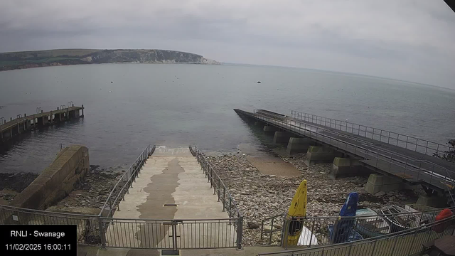 A view of a calm sea under a cloudy sky. In the foreground, there is a concrete pathway leading down to the water, bordered by metal railings. To the right, a ramp leads onto a larger platform extending into the water. Several colorful kayaks are lined up along the right side, sitting on the pebbly shore. In the background, gentle hills and cliffs can be seen in the distance.