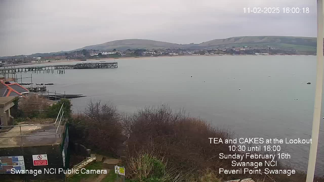 A gray, overcast sky looms over the shoreline of Swanage, with low hills in the background. The calm sea reflects the muted light, and a wooden pier extends into the water, flanked by a sandy beach. Lush greenery and shrubs partially obscure a sloping pathway leading down from a building in the foreground. Text overlays indicate an event for tea and cakes at the Lookout on Sunday, February 16th, along with the date and time.