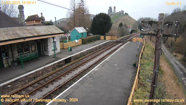 The image shows a railway station platform with several wooden benches and a small green shelter. To the left, there is a stone building with a sloped roof. In the background, there are trees and a hill with a castle or ruins at the top. The foreground features railway tracks that are lined with gravel, and a sign indicating "WAY OUT" is visible near the platform's edge. The sky is overcast, and the scene conveys a quiet, rural setting.