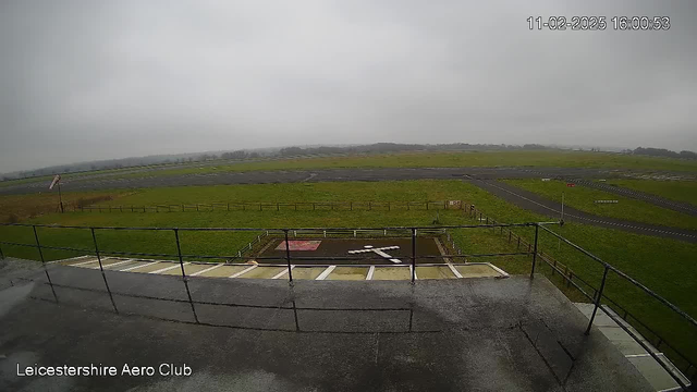 A cloudy and overcast sky is visible above a grassy airfield. In the foreground, there is a railing from a viewing platform. The airfield features a long, dark runway and a small area marked with a white cross, likely indicating a landing zone. A windsock is visible, indicating wind direction. Fenced grassy areas and a flat landscape extend into the background.