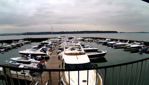 A cloudy view of a marina filled with various boats and yachts. The water is calm and reflects the gray sky. Numerous boats are moored in the dock area, with some parked closely together. In the foreground, a large white boat with a shelter is anchored, and wooden docks extend into the water. The marina appears to be surrounded by land in the background.