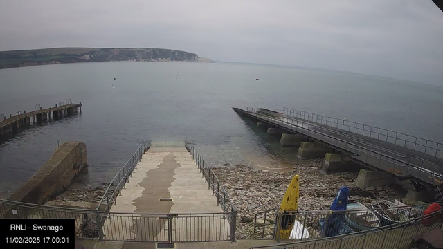 A view from a webcam showing a coastal landscape. In the foreground, a set of concrete steps leads down to rocky shorelines, with a few boats resting near the water's edge. A jetty extends out into the calm sea, and there are paddleboard and kayak storage in vibrant colors (yellow and blue) nearby. The backdrop features a cliffside with patches of greenery, under an overcast sky.