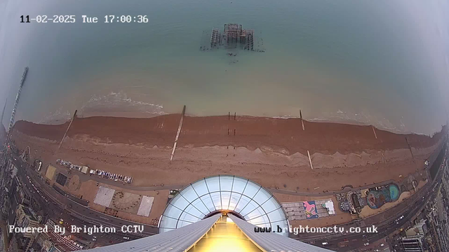 An aerial view from a webcam shows a sandy beach with a brown shoreline meeting a calm sea. In the middle of the image, the remnants of a pier are partially submerged in the water. The beach is lined with buildings and attractions, including a circular structure and a pool towards the right. The sky above is cloudy, and the time is displayed as 17:00:36 on February 11, 2025. A logo for Brighton CCTV appears in the bottom left corner.