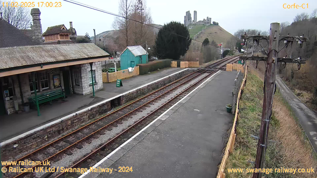 A view of a train station platform with an old stone building to the left. There is a green bench on the platform and a wooden fence in the background. Beyond the station, there are railway tracks that lead off to the right. In the distance, a hill rises, topped with the ruins of a castle. There are some bare trees and shrubs along the path of the railway, and a power pole stands on the right side of the image. The sky is overcast.