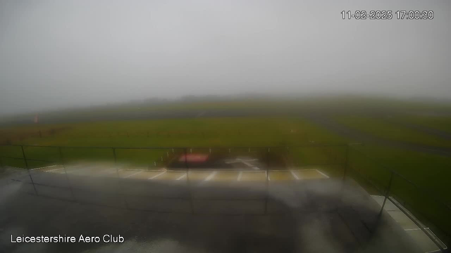 A foggy scene is depicted from a webcam at Leicestershire Aero Club, showing a blurred view of the runway and surrounding grass. The visibility is poor due to thick mist, making details indistinct. In the foreground, there is a safety fence and a portion of the tarmac, with a few blurred markings visible. The image is timestamped.