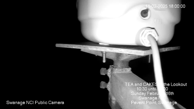 A black and white webcam image showing a metallic surface with a white container on top. The container has a visible pipe connected to it. Below the container, there are screws securing it to the surface. The background is dark, suggesting low-light conditions. Overlaid text provides event information, including "TEA and CAKES at the Lookout," with a date and time. The camera label is present in the bottom left corner.