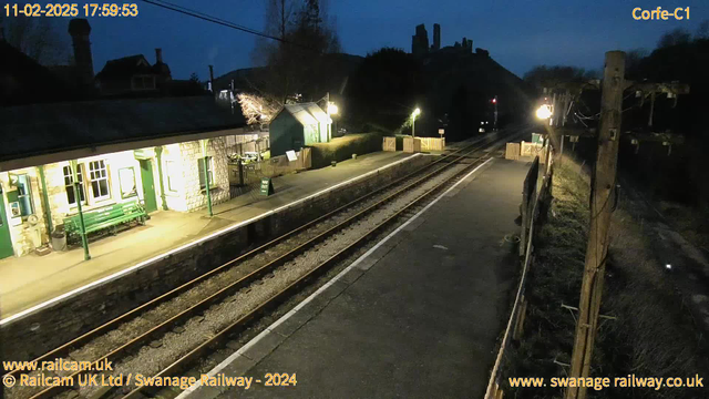 The image shows a train station at twilight. The platform is empty with a stone building featuring large windows on the left, and a green bench beside it. In the background, there are trees and a faint outline of hilltops. The tracks run alongside the platform, with wooden fencing visible on the right. A sign indicating "WAY OUT" is placed on the ground. Streetlights provide dim illumination, creating a tranquil evening atmosphere.