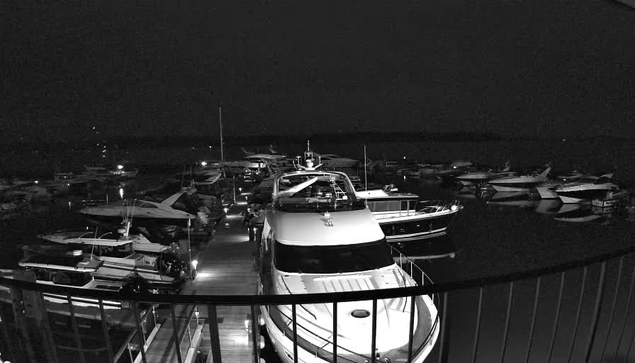 A nighttime scene at a marina showing several boats docked in the water. In the foreground is a large boat with a white hull and a black stripe, facing towards the viewer. Surrounding it are various other boats, some with noticeable shapes like cabin cruisers and smaller vessels. The docks are wooden, providing a path between the boats. The image is predominantly dark with faint reflections of the boats on the water's surface, suggesting a calm and quiet atmosphere.