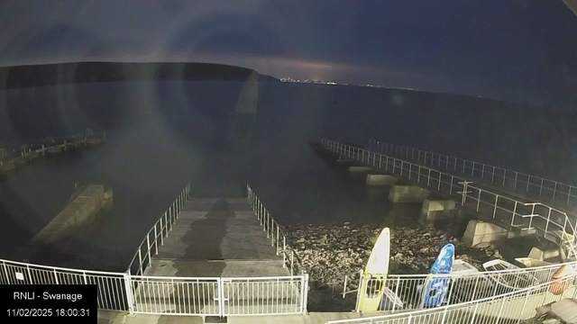 A dark shoreline scene with a calm sea under night sky. In the foreground, a wooden dock extends over rocky terrain, protected by white railings. To the right, two brightly colored kayaks are resting on the shore, one yellow and one blue. The background features a distant coastline illuminated by city lights, with a hilly silhouette. The image has a timestamp indicating the date and time at the bottom left corner.