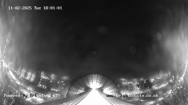 A black and white image from a webcam showing a view of a city at night. The scene includes a wide, illuminated pathway that leads toward a building or structure, with various lights visible on both sides, suggesting a busy urban area. The upper part of the image is darker, indicating a nighttime setting, while the timestamp in the corner shows "11-02-2025, Tue 18:01:01". The bottom left corner displays "Powered By Brighton CCTV" and the website "www.brightoncctv.co.uk".