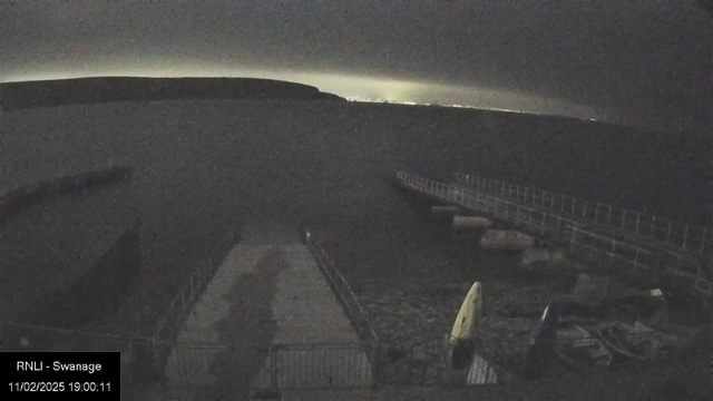 A dimly lit scene of a harbor at night. In the foreground, a dock extends over still water, with a railing along its edge. There are several rocks and structures near the shoreline. A small yellow kayak is visible on the dock. The horizon is faintly illuminated by distant lights, with a dark, cloudy sky above.