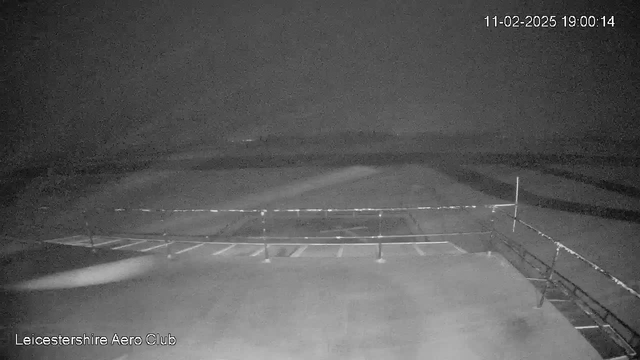 A dark, grainy image captured by a webcam shows an empty parking area at an aerodrome. In the foreground, a railing runs along the edge of the walkway, and several outlined parking spaces are visible. The background is mostly indistinct with some blurred lines suggesting runways or fields. The time and date are displayed in the upper right corner, indicating it's 7:00 PM on February 11, 2025.