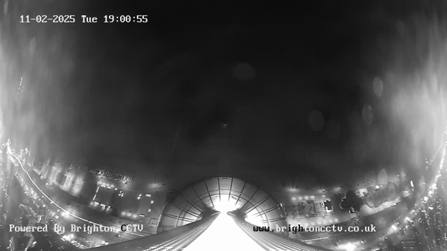 A black and white image from a webcam showing a view of a city street and buildings at night. The foreground features a downward slope leading away from the camera, with lights from vehicles and buildings visible along the sides, creating a sense of motion. The top of the image is mostly dark, with a timestamp in the upper left corner indicating the date and time. A logo for Brighton CCTV appears in the bottom left corner, and the website is displayed in the bottom right corner.