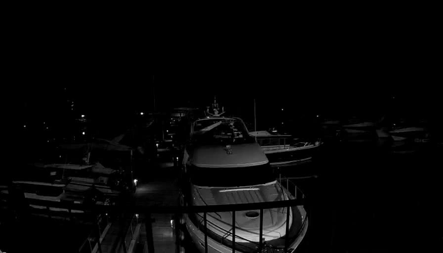 A dimly lit marina scene at night, featuring several boats docked closely together. The foreground shows the silhouette of a larger boat with a smooth, curved hull. In the background, additional boats are visible as dark shapes against a mostly black setting, with faint lights hinting at their outlines. The overall atmosphere is quiet and tranquil, with shadows dominating the scene.