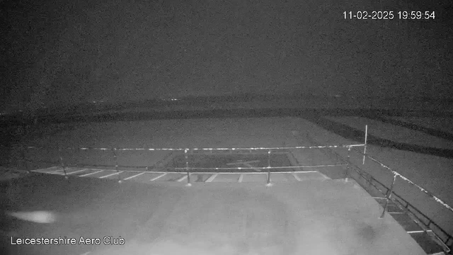 A dimly lit view from a webcam at night, showing a flat surface likely related to an airfield. There is a barrier or railing in the foreground. The background appears dark with some indistinct lights in the distance, indicating a nighttime setting. The date and time displayed at the top right corner read "11-02-2025 19:59:54." The bottom left corner indicates "Leicestershire Aero Club."