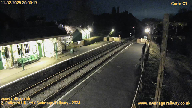 A dark image of a railway station at night. The scene includes a platform with a stone building that has a green roof and walls. There is a bench on the platform and a wooden fence in the background. A green sign labeled "WAY OUT" is visible. Train tracks lead off into the distance, with a light source illuminating part of the platform. In the background, there are trees and a silhouette of a hill.