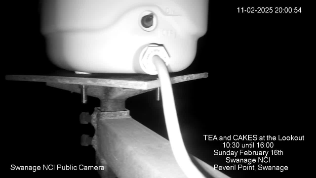 The image shows a close-up view of a white container with a spout, resting on a rectangular metal platform. A white cable is plugged into the bottom of the container. The background is dark, and in the bottom left corner, there is text indicating it is from the Swanage NCI Public Camera. Additional text on the right promotes a tea and cakes event, listing the date and time.