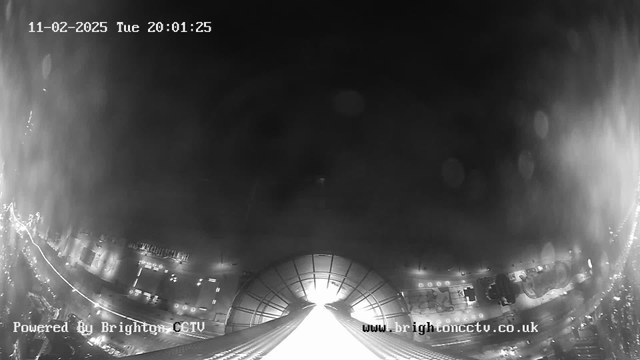 A black and white image captured by a webcam, showing a view looking down from a height. The bottom of the image features a bright, illuminated area suggesting a street or roadway, with faint outlines of buildings and vehicles visible. The upper portion is darker, with a soft gradient, indicating a night scene. The date and time are displayed in the upper left corner. A watermark for "Powered By Brighton CCTV" appears in the lower left, and a website URL is shown in the lower right corner.