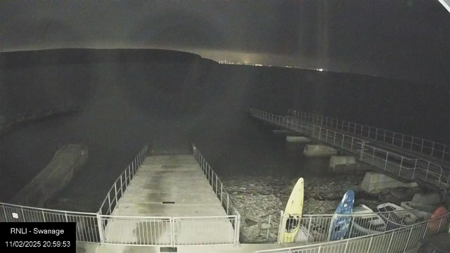 A dimly lit scene shows a harbor with a concrete pier extending into the water. The surface of the water appears dark and calm. On the left side of the image, a large rock is partially submerged. Two kayaks, one yellow and one blue, are resting on the pier. In the background, a faint outline of land can be seen against a night sky, with some distant lights suggesting the presence of a city. The foreground features a metal railing surrounding the pier, and the image has a timestamp in the lower left corner.