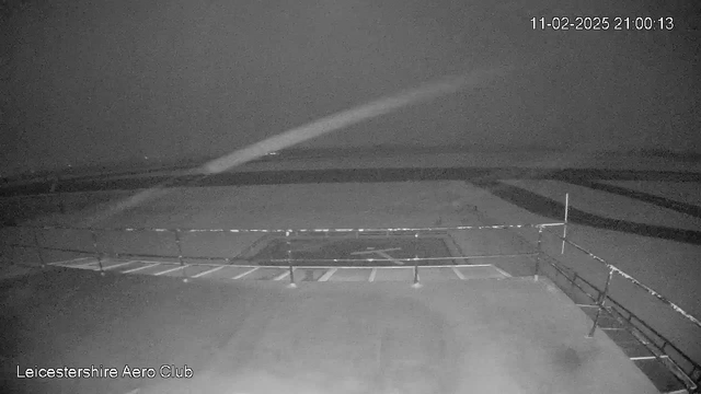 A dimly lit scene from a webcam at the Leicestershire Aero Club, showing an empty airstrip with a low-visibility horizon. A railing is visible in the foreground, and there are markings on the ground that indicate taxiways. The image has a grayish tint, suggesting nighttime conditions. The date and time are displayed in the top right corner.