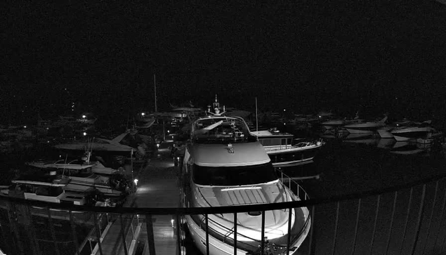 A dark, nighttime scene showing several boats docked at a marina. In the foreground, a large white yacht is prominent, with a partially visible deck and railing. Surrounding the yacht, other boats are moored in the water, which appears calm. A few small lights are visible, illuminating parts of the boats and the dock, creating a subtle contrast against the dark background. The overall atmosphere is tranquil and serene, typical of a quiet evening by the water.