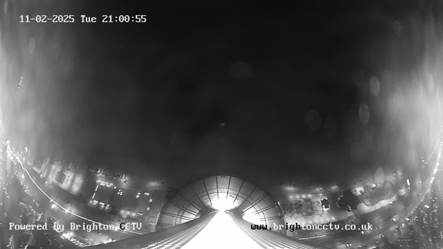 A dark, night-time scene captured from a webcam, showing a view of a city street. There are bright lights along the street, possibly from vehicles or street lamps, creating a contrast against the darkness. The corners of the image are blurred, and there are some light reflections or drops, indicating moisture on the lens. At the top of the image, the date and time are displayed: "11-02-2025 Tue 21:00:55." The bottom of the image includes a watermark that says "Powered By Brighton CCTV" and a website link.