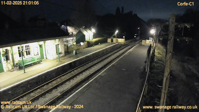 A dimly lit railway station at night. On the left, a platform with a green bench and stone walls. The platform has a small building with multiple windows, illuminated by a warm light. To the right, a fence with a "WAY OUT" sign. Train tracks stretch away into the distance, with a slight curve, and poles with lights alongside the tracks. The scene is quiet, with no visible train.