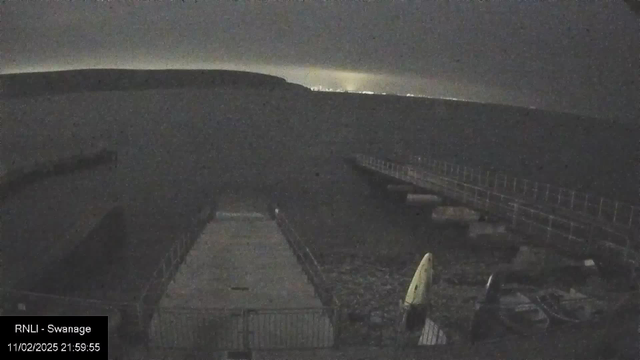 A dark scene shows a coastline with a pier extending into the water. In the foreground, there is a flat, concrete platform with a few scattered objects, possibly boats or equipment. To the right, a small yellow object, likely a kayak, is resting on the ground. The lighting is dim, revealing a silhouette of distant hills and a faint glow from a city or lights on the horizon. The overall atmosphere is calm and quiet, representing a nighttime setting by the sea.