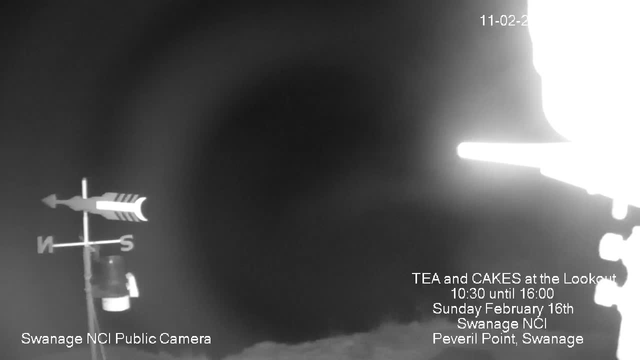 A black and white image from a webcam showing a dark scene with a partially obscured view. In the foreground, there is a weather vane indicating direction, with the letters "N" for North and "S" for South visible. To the right side of the image, there is bright white light that likely indicates an overexposed area. The bottom of the image includes text promoting a tea and cake event at the Lookout in Swanage, along with the date and location information. The overall ambiance is moody and shadowy, lacking significant detail.