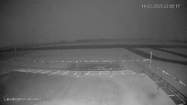 A dimly lit view from a webcam at the Leicestershire Aero Club shows a section of an airstrip. The foreground features a railing and a marked area on the ground, possibly a taxiway or runway. In the background, the airstrip extends into the distance, obscured by low visibility conditions. The timestamp indicates it is night.