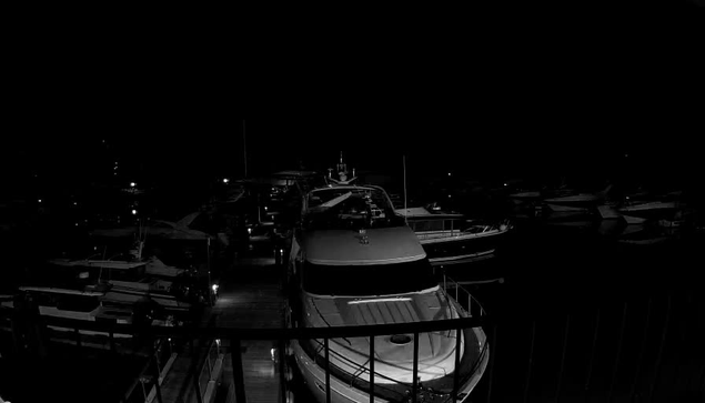 A dark scene featuring several boats docked at a marina. The image is primarily black and white, with some dim lights illuminating parts of the boats. A larger boat is positioned in the foreground, facing away from the viewer, while smaller boats are scattered in the background, creating a sense of depth. The water appears calm, reflecting the faint light.