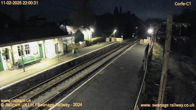 A dark scene at a railway station during night time. The platform has a few wooden benches and an information sign. On the left, there's a stone building that appears to be the station house with windows illuminated. A path and a small fence lead away from the platform. The railway tracks are visible running through the center, with faint lights reflecting off them. A wooden utility pole is on the right side of the image, and in the background, there are blurred outlines of buildings or trees. The overall atmosphere is quiet and tranquil.