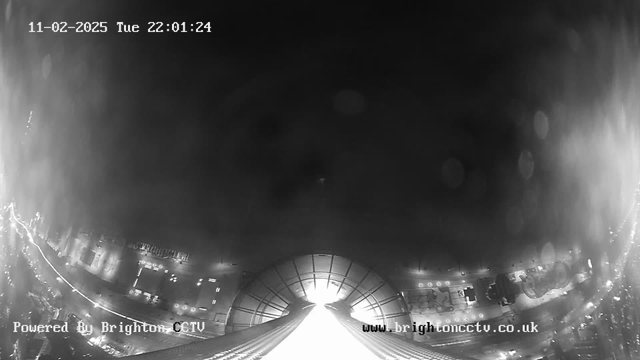 A dimly lit scene captured by a webcam, showing a wide view from a height. The foreground features a large, illuminated structure resembling a building or roof, leading into a bright, light-filled area. Below, faint outlines of streets and buildings can be seen, with scattered lights indicating vehicles or street lamps. The overall image is in black and white with soft shadows and a slight haze, indicating nighttime conditions. The timestamp at the top shows "11-02-2025, Tue 22:01:24," and there are water droplets appearing on the lens, creating a blurry effect. There are also text overlays indicating the camera is powered by Brighton CCTV.