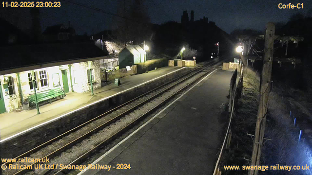 A nighttime image of a train station platform. The platform has a light green stone building with large windows and a green bench underneath them. There is a sign with "WAY OUT" visible on the ground near wooden fencing. To the right, there are two railway tracks with gravel in between. A wooden utility pole with cables stands at the edge of the image, and faint illumination highlights the surrounding area, creating a somewhat dim atmosphere.