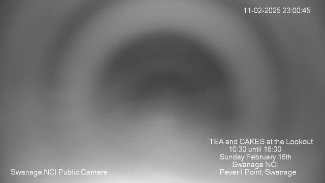 A blurred grayscale image, possibly taken from a webcam. The central area appears darker, creating a circular gradient effect. Text in the lower left corner reads "Swanage NCI Public Camera," while text in the lower center details an event: "TEA and CAKES at the Lookout, 10:30 until 16:00, Sunday February 16th, Swanage NCI, Peveril Point, Swanage." In the upper right corner, there is a date and time stamp: "11-02-2025 23:00:45." The overall appearance is indistinct and abstract.