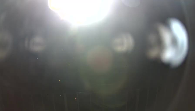 A bright light from the top center dominates the image, creating glare and making it difficult to see details. The foreground appears blurry with indistinct shapes, possibly from the camera lens. The background is dark, suggesting a lack of visible features or context.